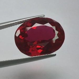 May include: A deep red, oval-shaped gemstone with a faceted surface that reflects light. The stone is held by a pair of silver-toned metal tweezers with a textured grip pattern on the handle, showcasing the clarity and vibrant colour of the gem.