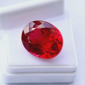 Peut inclure: Une pierre précieuse ovale d'un rouge vif avec une surface à facettes qui reflète la lumière. La pierre précieuse est présentée à l'intérieur d'une boîte à bijoux rectangulaire blanche dotée d'une doublure intérieure douce et rembourrée.