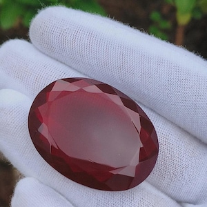 May include: A large, oval-cut, deep red gemstone. The gem is held in a white fabric glove, with the facets reflecting light. The gemstone's colour is a rich, dark red, with a polished surface. The overall shape is elongated, with a symmetrical design.