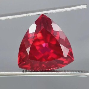 以下が含まれることがあります： 三角形にカットされたルビーの宝石。宝石は鮮やかな赤色で、灰色の背景に対してピンセットで保持されています。石のカットは、複数の反射面を作り出しています。