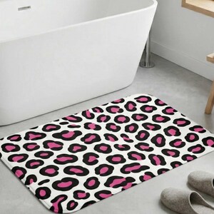 May include: A rectangular bath mat with a white background and a pink and black leopard print pattern. The mat is placed in front of a white bathtub. The bathroom has a neutral color scheme.