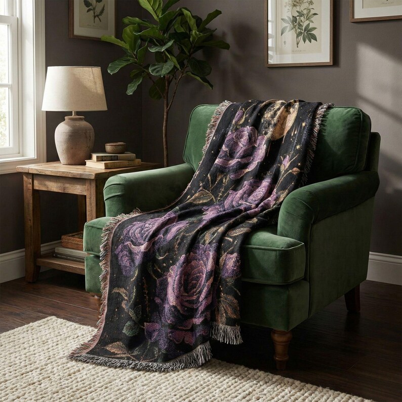 May include: A black throw blanket with a floral rose design in shades of purple and gold is draped over a green armchair. The blanket has a fringed edge and is placed in a cozy living room setting.