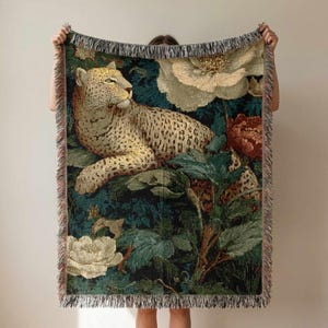 May include: Woven throw blanket featuring a leopard resting amidst lush foliage and flowers in shades of green, cream, and red. The fringed edges add a decorative touch to this home decor item.