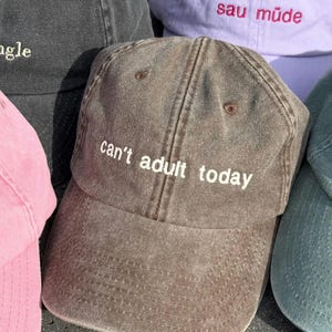 Könnte beinhalten: Eine braune Baseballkappe mit dem in weißem Garn gestickten Text can't adult today. Die Kappe hat ein verwaschenes, abgenutztes Finish und ist von weiteren Baseballkappen in den Farben Schwarz, Pink, Helllila und Dunkelgrün umgeben.