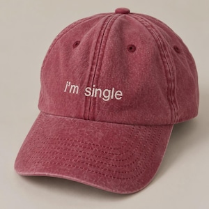 May include: A washed-out burgundy baseball cap with a six-panel construction and a button on top. The front features the embroidered text "I'm single" in white thread. The cap has a curved brim with visible stitching and matching eyelets.