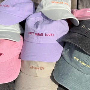 Könnte beinhalten: Eine Sammlung von Baseballkappen in verschiedenen Pastell- und gedeckten Farben wie Lavendel, Rosa, Beige, Weiß und Anthrazit. Jede Kappe ist auf der Vorderseite mit Text bestickt, wie zum Beispiel 'can't adult today', 'sau müde', 'Growth', 'I'm single' und 'wird schon passen'.