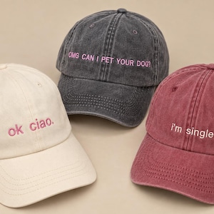 Könnte beinhalten: Drei Baseballkappen mit gesticktem Text. Eine cremefarbene Kappe zeigt den rosa Schriftzug 'ok ciao.', eine anthrazitfarbene Kappe den rosa Schriftzug 'OMG CAN I PET YOUR DOG?' und eine kastanienbraune Kappe den weißen Schriftzug 'i'm single'.
