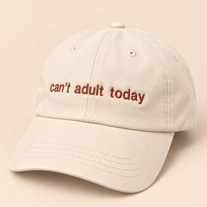 Op de afbeelding: Een beige honkbalpet met de geborduurde tekst can't adult today in donkerbruine draad. De pet heeft een gestructureerde kroon, een gebogen klep en gestikte oogjes op de bovenste panelen, wat zorgt voor een strak en eenvoudig ontwerp.