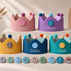 Könnte beinhalten: Fünf Stoffkronen in Rosa, Lila, Gelb, Dunkelblau und Türkis, jeweils mit Pompons und einem runden Abzeichen mit der Zahl 1 verziert. Die Kronen sind mit den Namen Marie, Emma, Finn, Emilio und Emil bestickt. Zehn weitere nummerierte Abzeichen liegen davor.