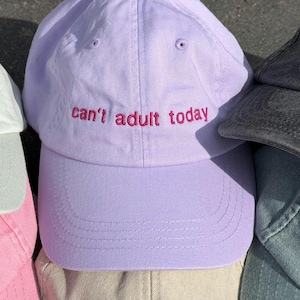 Könnte beinhalten: Eine Sammlung von Baseballkappen in verschiedenen Farben, darunter Weiß, Hellrosa, Lavendel, Beige, Dunkelgrau und gedecktes Grün. Die lavendelfarbene Kappe zeigt den gestickten Text 'can't adult today' in Pink, die dunkelgraue zeigt 'I'm sing'.