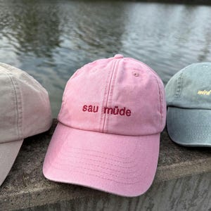 Può includere: Un cappellino da baseball rosa con visiera curva e struttura a sei pannelli. La parte anteriore presenta il testo ricamato "sau müde" in filo rosso scuro. Il cappellino ha un bottone sulla parte superiore e cuciture visibili.