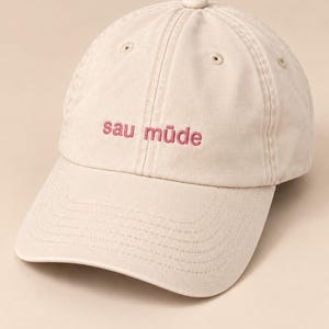 Könnte beinhalten: Eine beige Baseballkappe mit dem gestickten Text "sau müde" in dunkelrosa Garn. Die Kappe hat eine Sechs-Panel-Konstruktion mit einem gebogenen Schirm, einem stoffbezogenen Knopf an der Oberseite und genähten Ösen zur Belüftung an jedem Panel.