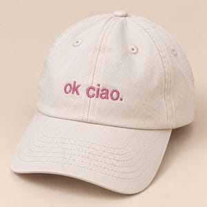 Puede incluir: Una gorra de béisbol de color beige con el texto "ok ciao." bordado en hilo rosa oscuro. La gorra tiene una corona estructurada con ojales de ventilación y una visera curva con detalles de costuras visibles en la parte frontal.