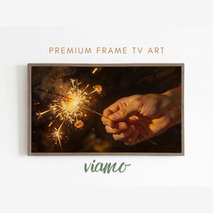 Pode incluir: Uma impressão de arte emoldurada com uma mão segurando uma faísca acesa, com faíscas brilhantes contra um fundo escuro. O texto "PREMIUM FRAME TV ART" está no topo e "viamo" na parte inferior.