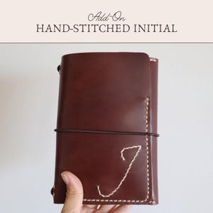Puede incluir: Una funda de cuaderno de cuero marrón oscuro con el texto Add-On HAND-STITCHED INITIAL en la parte superior. La funda se asegura con una banda elástica fina de color marrón oscuro y muestra una letra J grande cosida a mano en color crema.