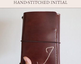 May include: A dark brown leather notebook cover featuring the text Add-On HAND-STITCHED INITIAL at the top. The cover is secured with a thin dark brown elastic band and displays a large, cream-colored hand-stitched letter J on the front panel.
