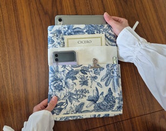 May include: A fabric sleeve featuring a blue and white floral toile pattern. The sleeve has a front pocket containing a small card with the text CICERO, a folded smartphone, a metal binder clip, and a pen. A tablet is inserted into the top.