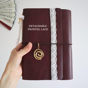 May include: A dark burgundy journal with a vertical white lace trim and a gold-toned spiral charm attached to a dark brown elastic band. The cover features the white text DETACHABLE FRONTAL LACE. The journal has a smooth, matte finish.