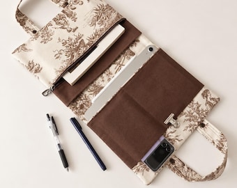 May include: A fabric organizer case featuring a cream and brown toile de Jouy print with two handles. The interior has brown fabric pockets holding a notebook, a tablet, a small folding smartphone, and a metal binder clip, with two pens nearby.