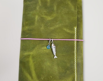 May include: A rectangular olive green leather journal cover featuring a distressed texture and white stitching along the edges. A thin pink elastic band wraps horizontally around the center, holding a silver fish charm and a small blue evil eye charm.