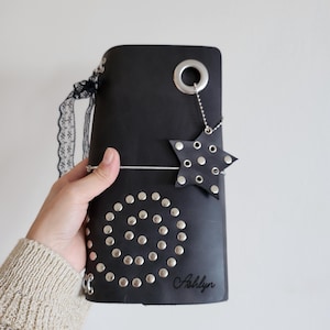 May include: A black leather journal cover featuring a spiral pattern of silver metal studs on the front. It includes a large silver eyelet at the top, a dangling star-shaped charm with studs, a black lace ribbon, and the name Ashlyn embossed in script.