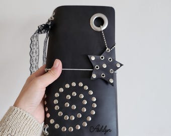 May include: A black leather journal cover featuring a spiral pattern of silver metal studs, a large silver eyelet at the top, and a star-shaped charm with studs attached by a chain. The name Ashlyn is embossed on the front, with black lace trim.