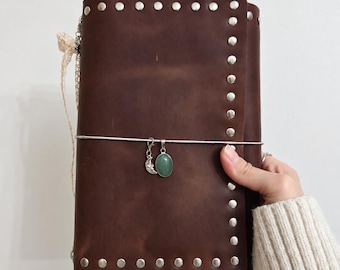May include: A dark brown leather journal cover featuring a vertical row of silver-toned metal studs along the right edge. A silver elastic band wraps around the center, holding a small silver crescent moon charm and an oval green stone pendant.
