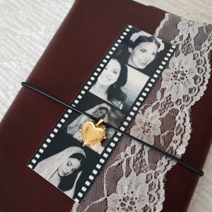 May include: A dark maroon journal wrapped with a white lace ribbon and a black elastic cord. A vertical film strip featuring four black and white portraits is tucked under the cord, accented by a small, polished gold heart-shaped pendant.