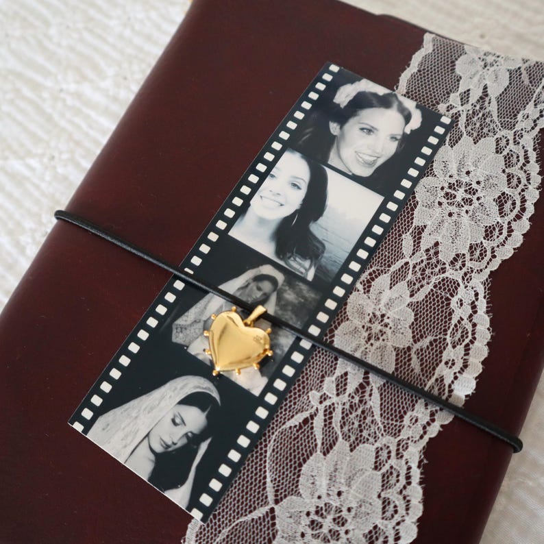 May include: A dark maroon journal wrapped with a white lace ribbon and a black elastic cord. A vertical film strip featuring four black and white portraits is tucked under the cord, accented by a small, polished gold heart-shaped pendant.