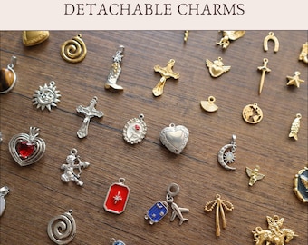 May include: A collection of assorted metal charms labeled Add-On DETACHABLE CHARMS. The charms include gold and silver-toned shapes such as a cowboy boot, crosses, a heart locket, a sun, a moon, a red enamel rectangle, a blue suitcase, and an airplane.