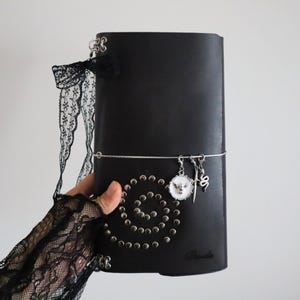 Könnte beinhalten: Ein schwarzes Lederjournal mit einem spiralförmigen Muster aus silbernen Metallnieten auf dem Einband. Das Journal ist mit einem schwarzen Spitzenband, einem silbernen Gummiband und drei Anhängern verziert: einem weißen runden Emblem, einem Schwert und einer Schlange.