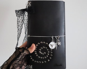 May include: A black leather journal featuring a spiral pattern of silver metal studs on the front cover. The journal is accented with a black lace ribbon, a silver elastic band, and three hanging charms: a white circular emblem, a sword, and a snake.