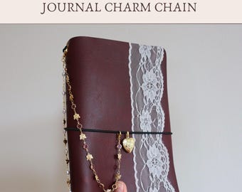 May include: A dark maroon journal featuring a vertical white lace trim and a black elastic band. Attached to the band is a gold-toned charm chain with small pink and clear beads, along with a heart-shaped locket. Text reads: Add-On JOURNAL CHARM CHAIN.