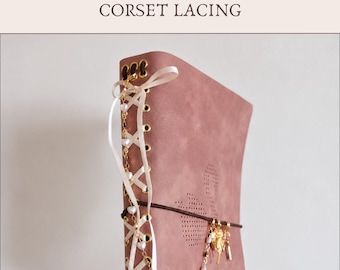 May include: A dusty rose journal featuring corset lacing with cream-colored ribbon threaded through gold-toned eyelets. A gold chain with small white heart beads hangs from the lacing, and a dark brown elastic band holds gold charms.