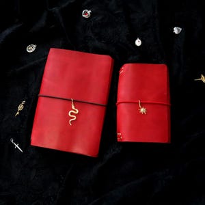 May include: Two red leather journals with elastic bands. The larger journal features a gold snake charm, while the smaller journal has a gold sun charm. Several small metal charms, including a key, a sword, and hearts, are scattered around.