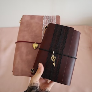 May include: Two rectangular journals with covers in dusty rose and dark burgundy. The rose journal features white lace trim and a gold heart charm. The burgundy journal has black lace trim and a gold key charm, both secured with elastic bands.