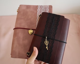 May include: Two journals with matte covers, one dusty rose and one dark burgundy. The rose journal features white lace trim and a gold heart locket charm. The burgundy journal features black lace trim and a gold key charm on an elastic band.