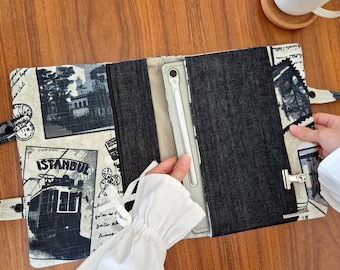 May include: A fabric tablet sleeve featuring a cream-colored background with black and white vintage-style prints of Istanbul, including a tram and postmarks. The sleeve has a dark denim center panel, a metal clip closure, and a light-colored stylus.