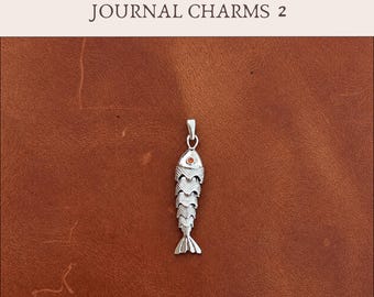 May include: A silver-toned fish-shaped charm pendant featuring a small orange stone eye and textured scale details. The text Add-On JOURNAL CHARMS 2 is displayed above the charm. The pendant includes a small metal loop for attachment.