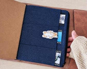 May include: A brown leather notebook cover featuring a dark blue fabric insert with three horizontal pockets. The pockets contain small rectangular photo prints and film strips. A small sticker of an orange tabby cat is attached to the center.