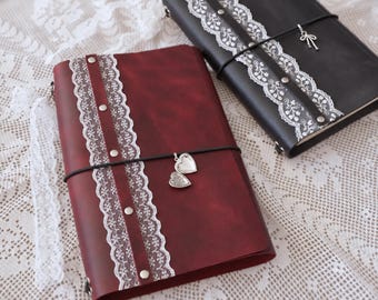 May include: A deep red journal and a black journal, each featuring a vertical strip of white lace secured with silver-toned rivets. The red journal has a silver heart-shaped locket charm, while the black journal has a silver bow charm.