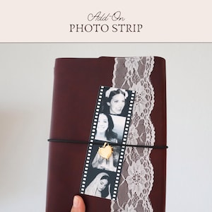 May include: A dark maroon leather-bound journal featuring a vertical strip of white floral lace and a black and white photo strip held in place by a thin black elastic band. A small gold heart-shaped charm is attached to the elastic over the photos.