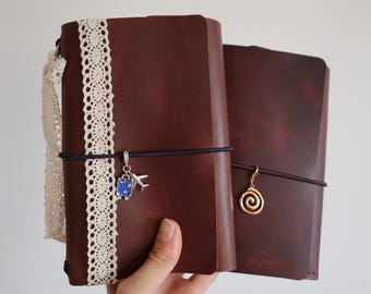 May include: Two dark brown leather journals. The left journal features a vertical cream-colored lace trim, a white lace bow, and a dark elastic band holding a blue suitcase and silver airplane charm. The right journal has a gold spiral charm.