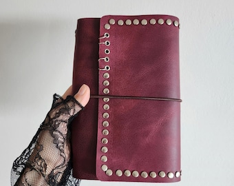 May include: A deep burgundy journal cover featuring a textured surface and a vertical row of silver-toned metal studs along the front edge. The cover is secured by a thin, dark brown elastic cord wrapped horizontally around the center.