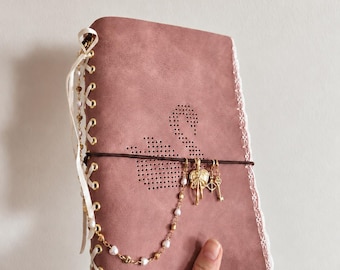 May include: A dusty rose journal featuring a perforated swan design on the cover. The spine is laced with white ribbon through gold eyelets. A dark brown elastic band holds a gold-toned charm chain with pearls, a bow, and a key.
