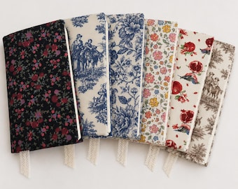 May include: Six rectangular fabric-covered notebooks arranged in a fan shape. The covers feature various patterns including dark floral, blue and white toile, large blue floral, small colorful flowers, pomegranate prints, and brown and white toile.