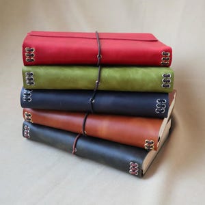 May include: A stack of five rectangular journals with smooth covers in red, olive green, navy blue, tan, and dark grey. Each journal features metal eyelets along the spine edges with thin cord lacing and a vertical elastic band closure.