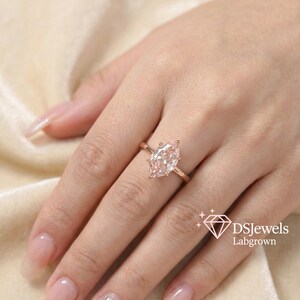 May include: A rose gold ring with a marquise-cut, lab-grown diamond. The ring is set on a delicate band and is displayed on a hand with manicured nails. The image includes the text "DSJewels Labgrown".