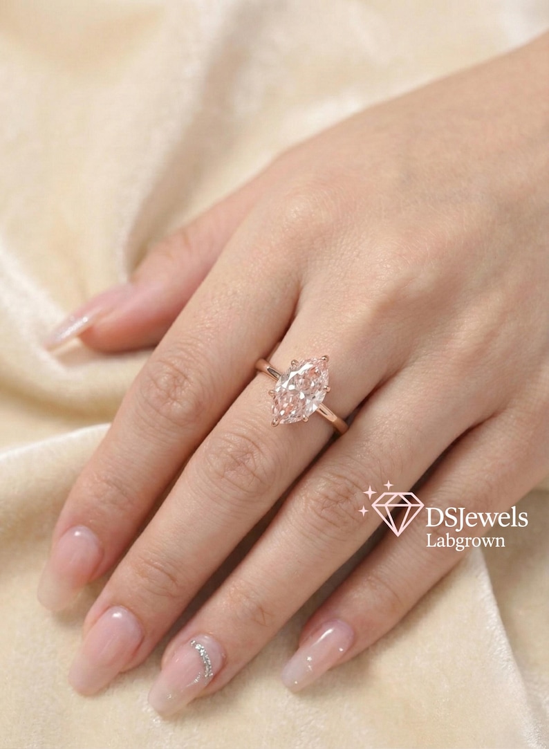 May include: A rose gold ring with a marquise-cut, lab-grown diamond. The ring is set on a delicate band and is displayed on a hand with manicured nails. The image includes the text "DSJewels Labgrown".