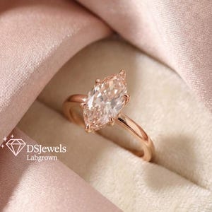 May include: A rose gold engagement ring with a marquise-cut lab-grown diamond. The diamond is a light peach color and set in a four-prong setting. The ring is displayed on a pink satin fabric. The text "DSJewels Labgrown" is visible.